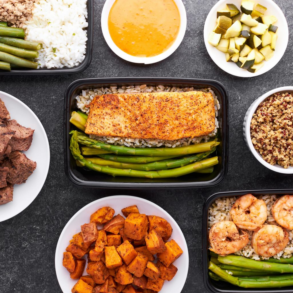 Flat lay of meal prep containers featuring salmon, asparagus, shrimp, and sweet potatoes.